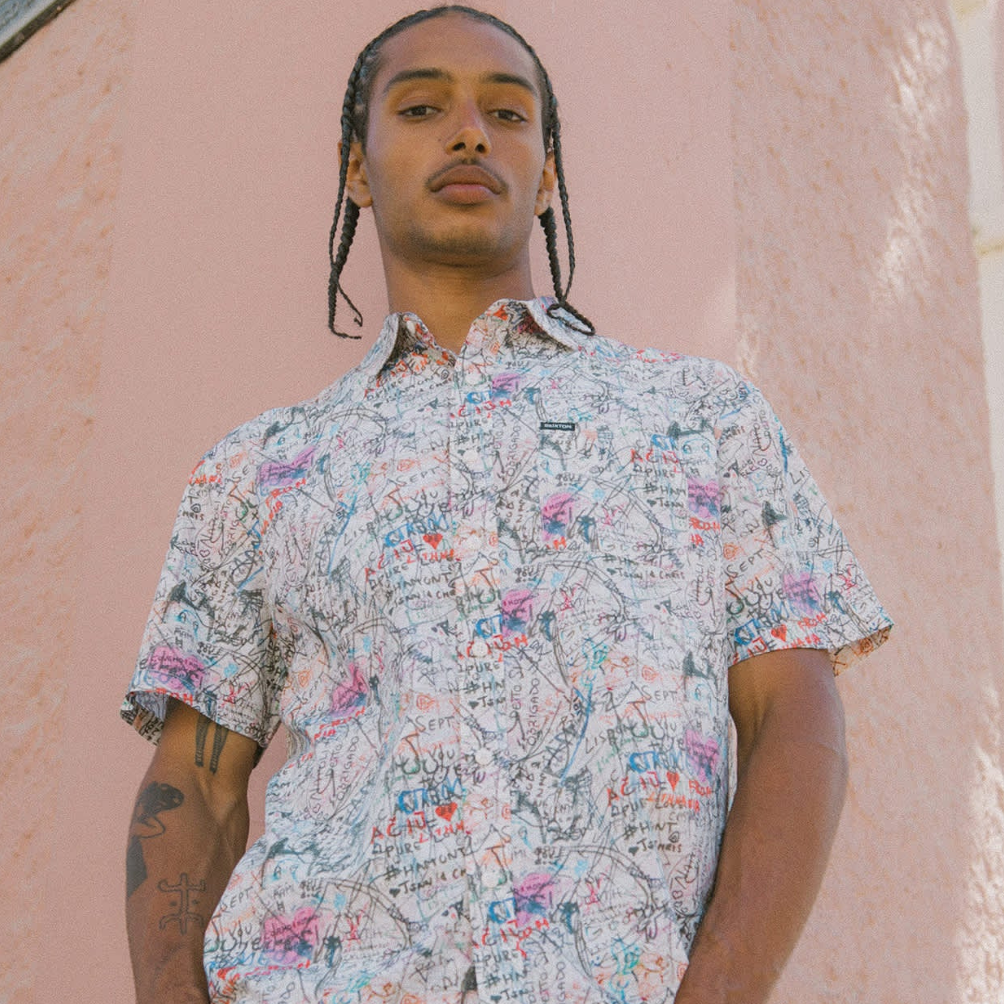 man wearing a brixton cru short sleeve woven shirt with graffiti pattern on a pink background