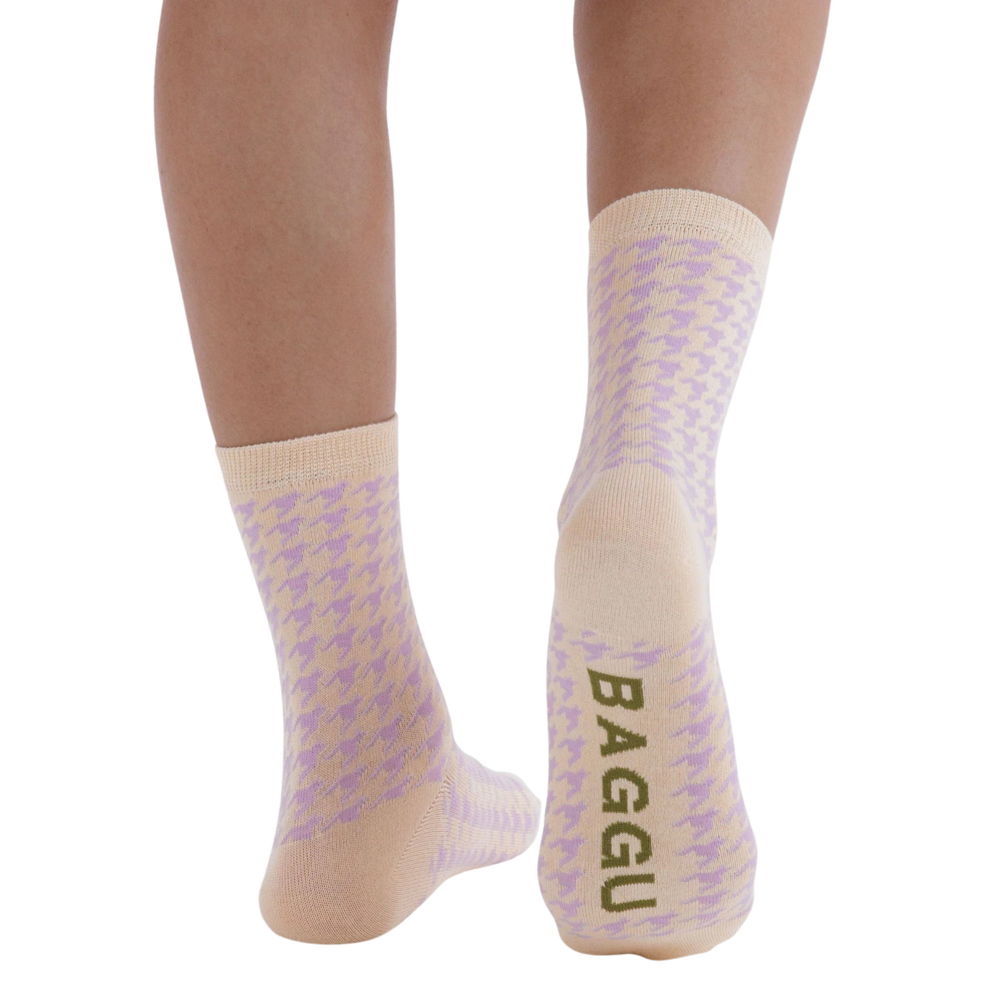 A pair of baggu crew socks with a light purple and beige houndstooth pattern, worn on a person's feet against a plain white background. The brand name 'BAGGU' is printed vertically in olive green on the sole of the right sock.