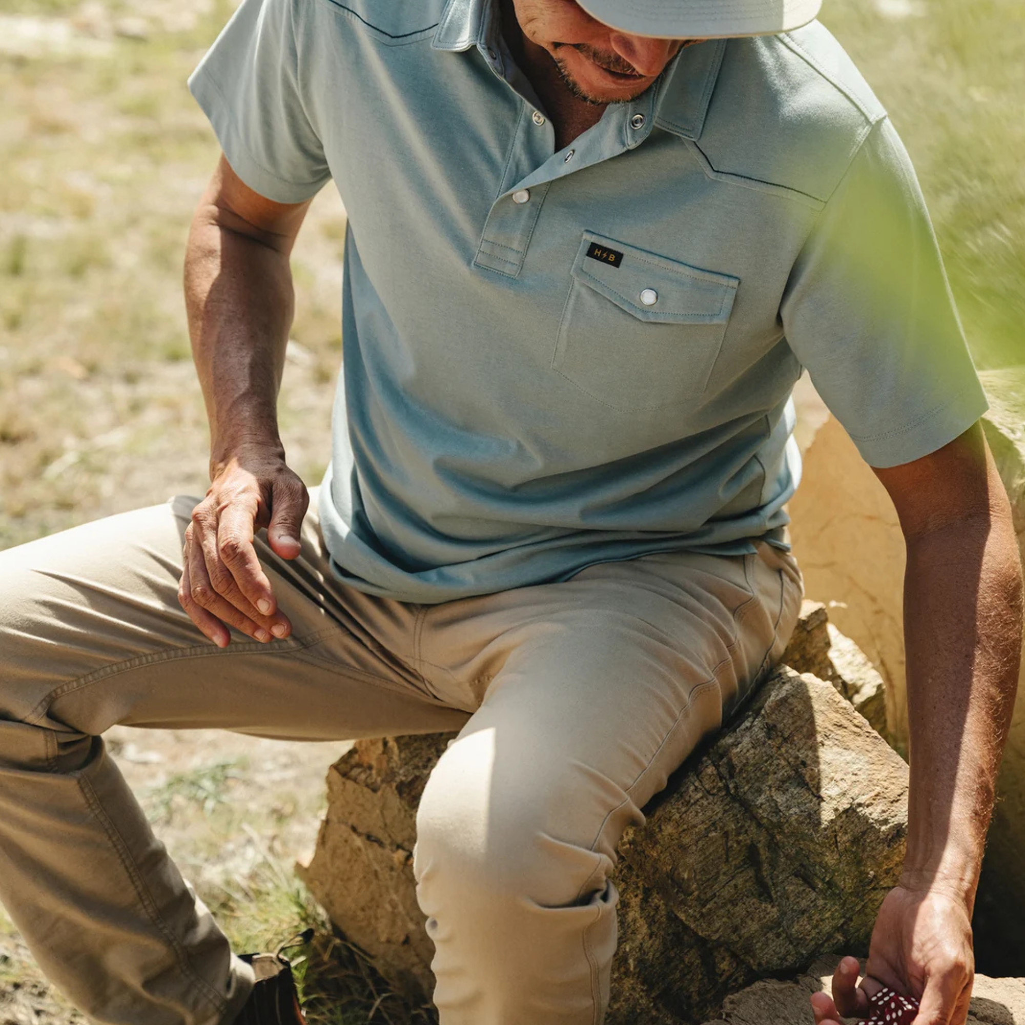 A man wearing a light blue short-sleeve ranchero polo shirt and khaki pants sits outdoors on a rock in a sunny, grassy environment.