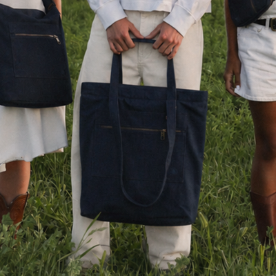 A navy blue canvas tote bag with double shoulder straps and a front zip pocket, held by a person standing in a grassy outdoor setting. Other people are partially visible on both sides.