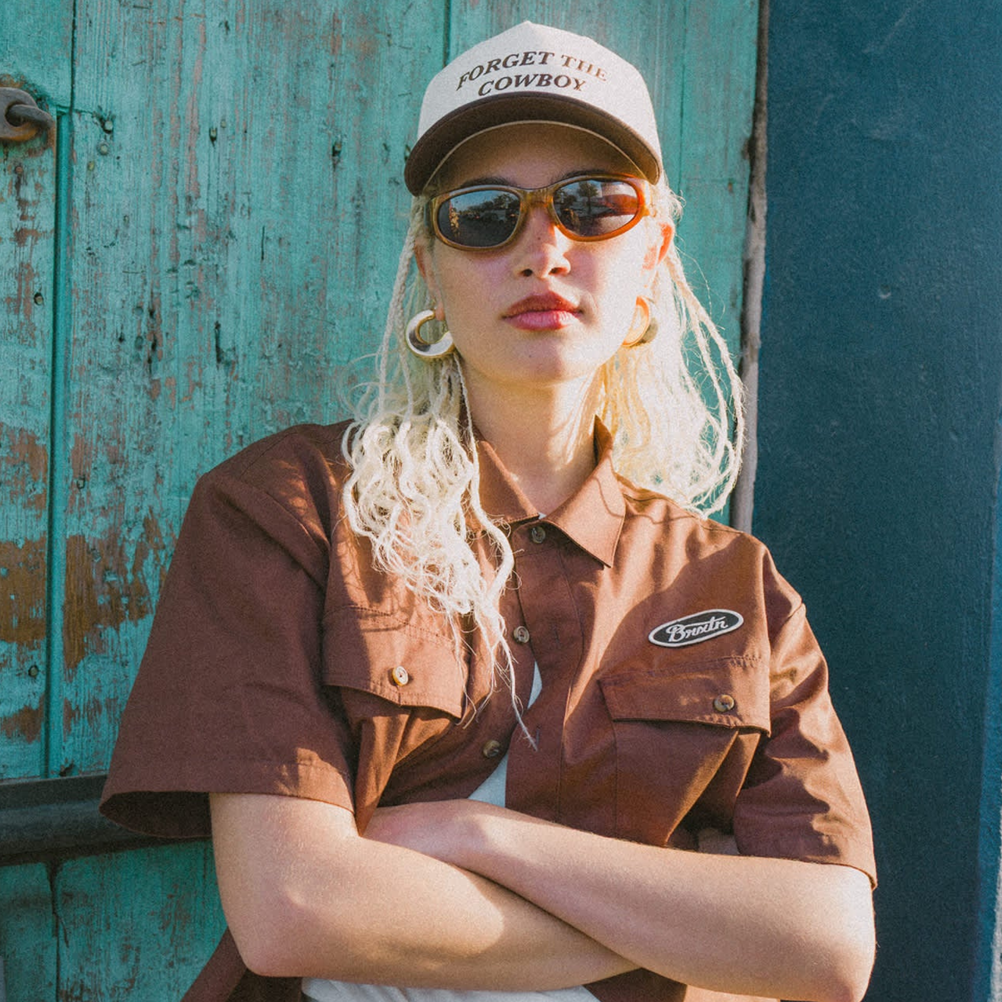 girl wearing a white and brown brixton hat with forget the cowboy in brown stitching on a blue background