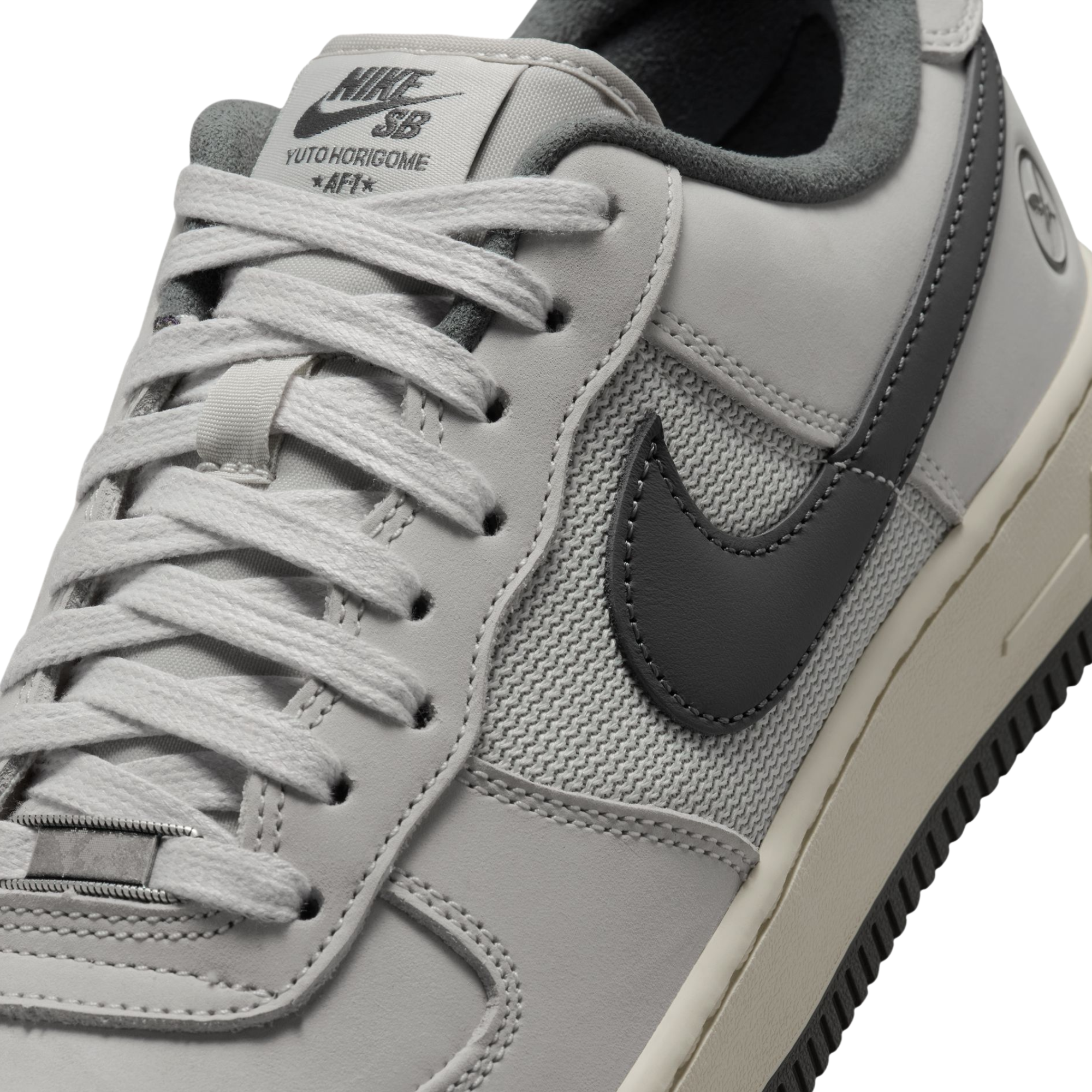 A close-up of a Nike SB Yuto Hoshime AF1 sneaker in light gray and dark gray, showing the side panel, laces, and Nike branding.