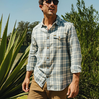 A man wearing a light blue and beige plaid button-down shirt with brown buttons, styled with khaki pants and sunglasses, standing outdoors in front of green plants under a clear blue sky.