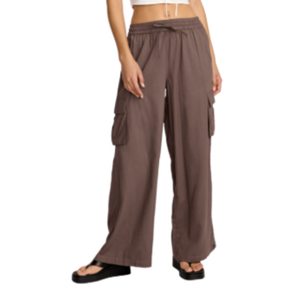 Woman wearing a white crop top and brown wide-leg volcom lived in beach pants on a white background