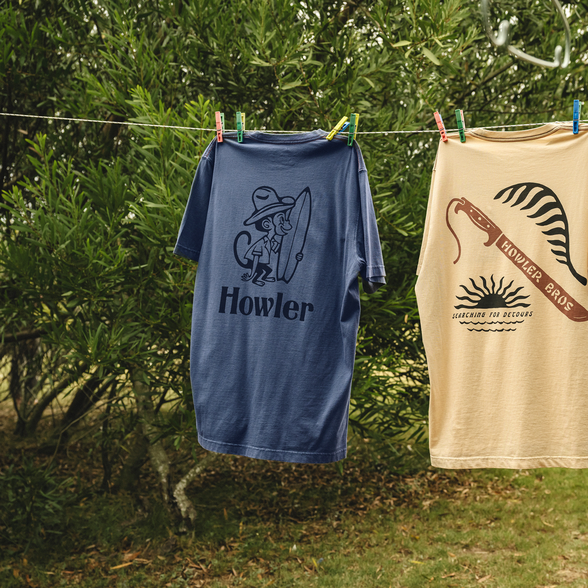 Two graphic t-shirts hanging on a clothesline outdoors in front of green bushes. The left shirt is navy blue with a dark blue graphic of a surfer monkey and the text 'Howler'.